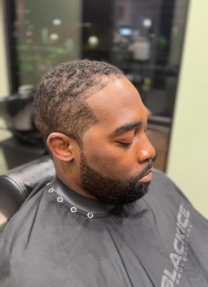 A close-up of a man with a fresh haircut and groomed beard, wearing a barber cape inside a modern barbershop.