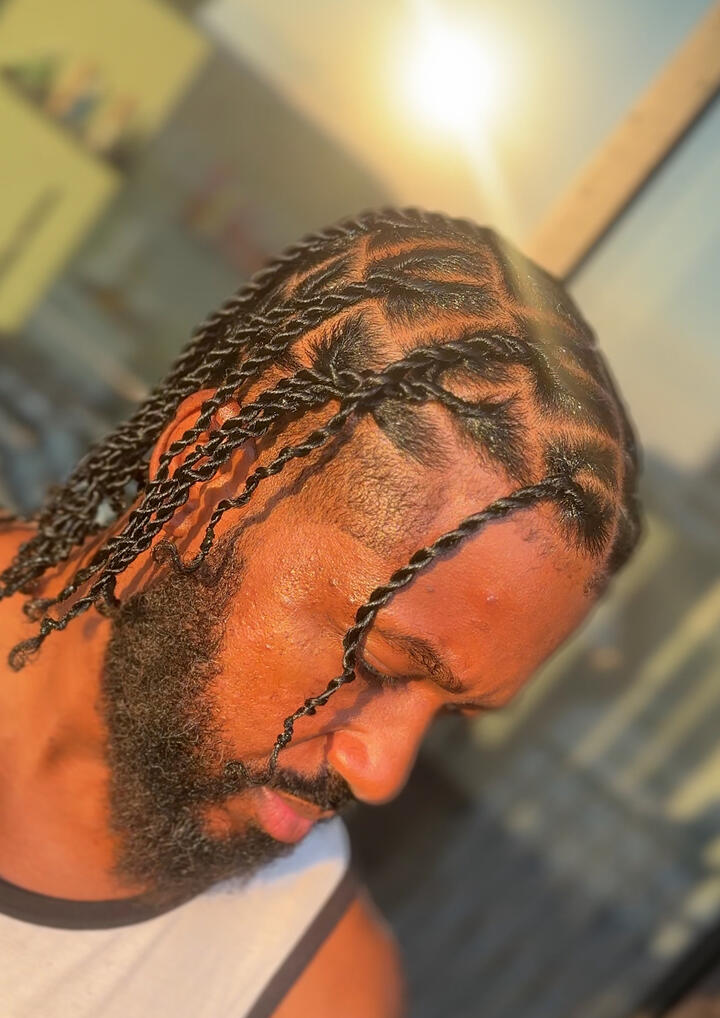 Man with a full beard and neatly twisted braids, captured in warm sunlight while looking downward.
