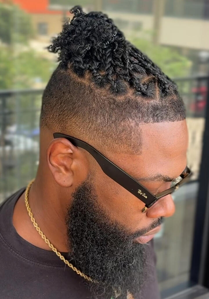 A close-up of a man with a fresh haircut and groomed beard, wearing a barber cape inside a modern barbershop.